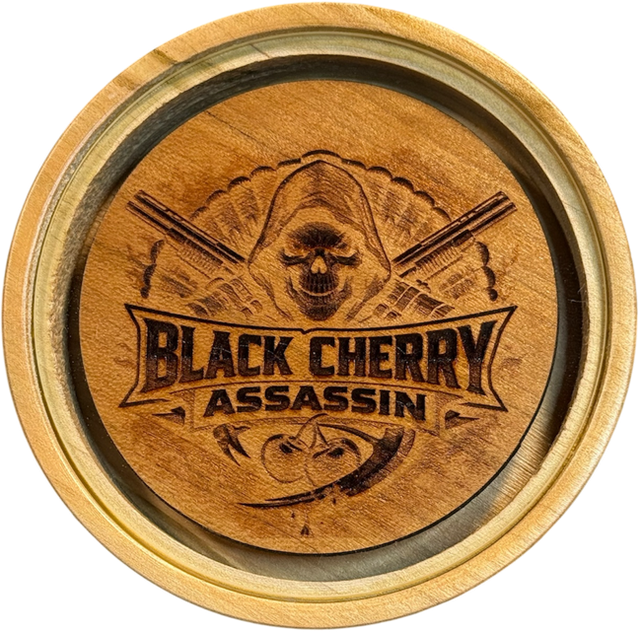 The Black Cherry Assassin pot call: a hand-turned solid cherry wood call with a laser-engraved design of a hooded skull and crossed turkey call strikers, and a crystal striking surface inset at the center.